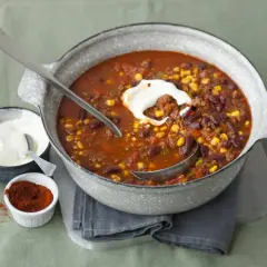 Herzhaftes Chili con Carne mit Bohnen, Mais und Hackfleisch, garniert mit einem Klecks Sauerrahm. Daneben eine Schale mit Sauerrahm und ein Schälchen mit Chilipulver.