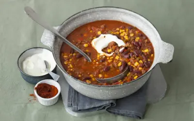 Herzhaftes Chili con Carne mit Bohnen, Mais und Hackfleisch, garniert mit einem Klecks Sauerrahm. Daneben eine Schale mit Sauerrahm und ein Schälchen mit Chilipulver.