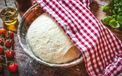 Frischer Teig für ein Pizzateig Rezept ruht in einer Schüssel, bedeckt mit einem rot-weiß karierten Tuch. Tomaten und Olivenöl liegen bereit.
