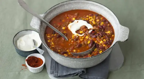 Herzhaftes Chili con Carne mit Bohnen, Mais und Hackfleisch, garniert mit einem Klecks Sauerrahm. Daneben eine Schale mit Sauerrahm und ein Schälchen mit Chilipulver.