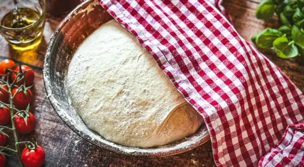 Frischer Teig für ein Pizzateig Rezept ruht in einer Schüssel, bedeckt mit einem rot-weiß karierten Tuch. Tomaten und Olivenöl liegen bereit.