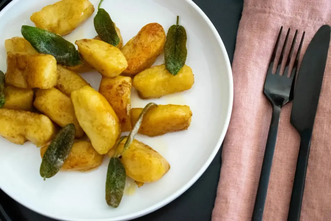 Goldbraune salbei gnocchi auf weißem Teller, garniert mit frischen Salbeiblättern. Daneben liegen schwarzes Besteck auf rosafarbenem Stoff.