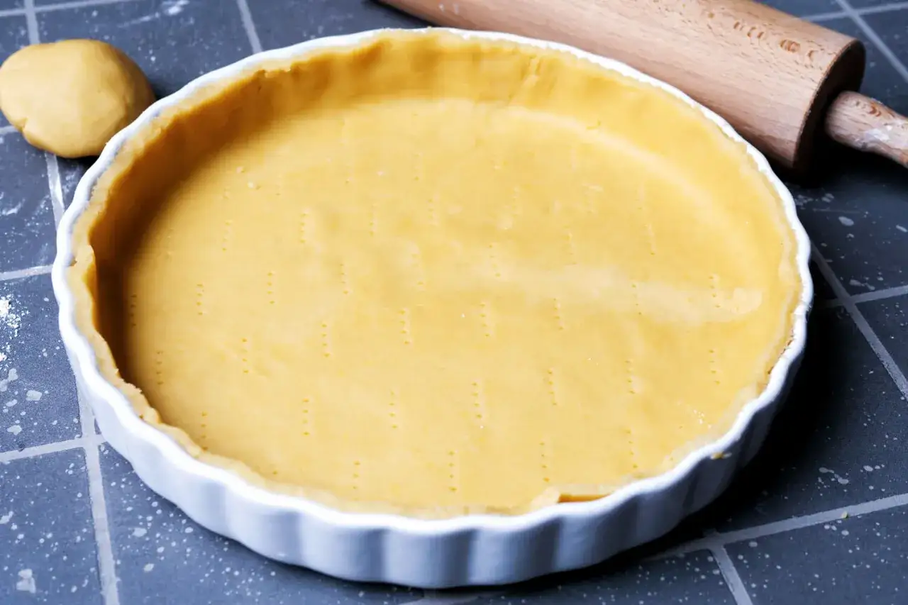 Ein vorbereiteter quiche teig liegt in einer wei&szlig;en Tarteform. Daneben ein Nudelholz und ein Teigball.