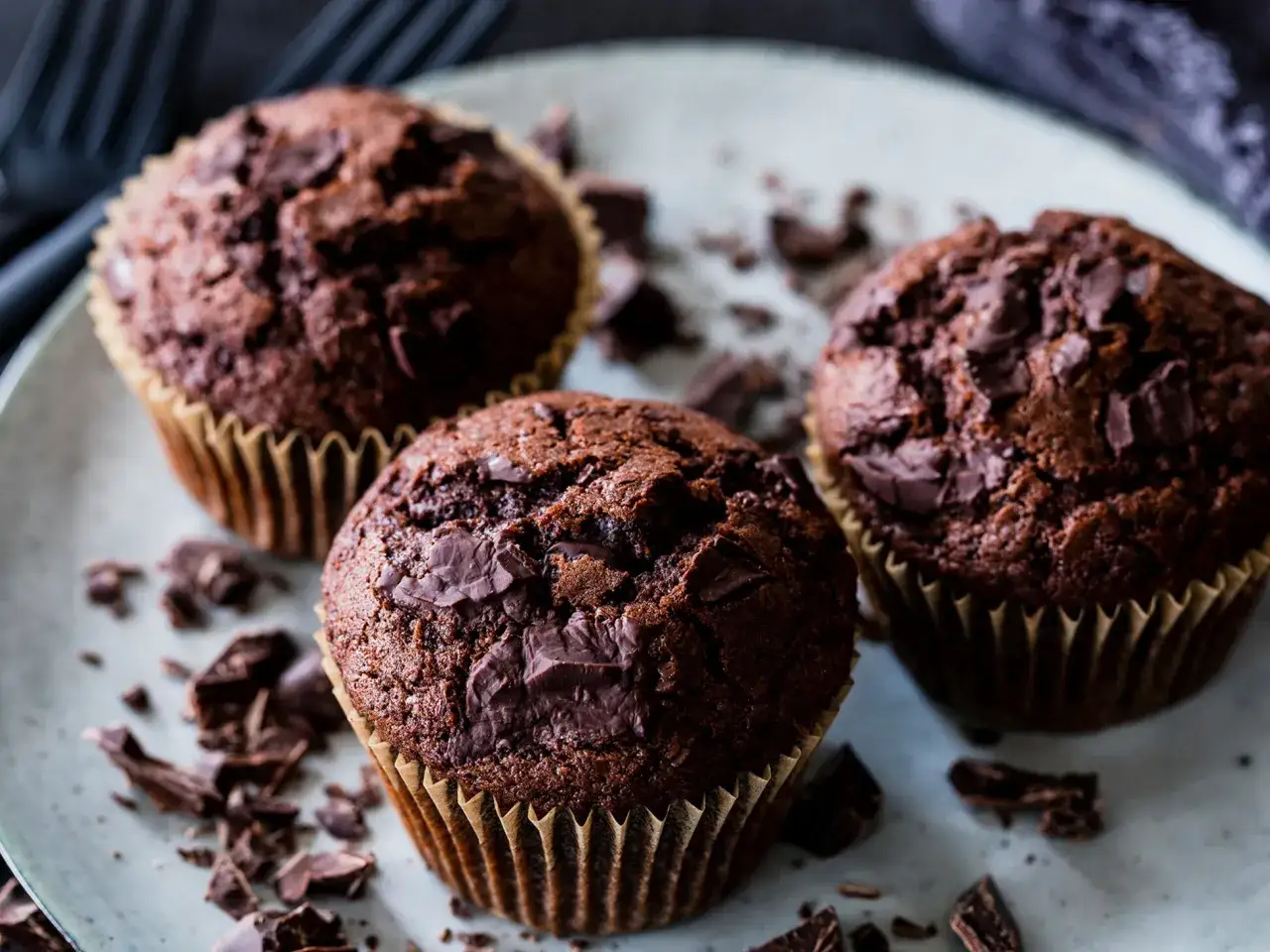 Drei saftige triple chocolate muffins mit Schokoladenstückchen auf einem Teller, umgeben von Schokoladenraspeln.