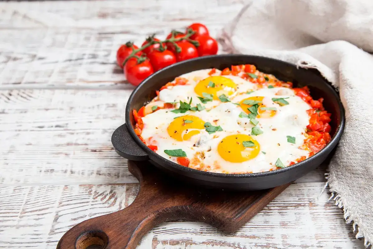 Ein k&ouml;stliches Shakshuka Rezept in einer Gusseisenpfanne, garniert mit Petersilie. Daneben ein Bund Kirschtomaten.