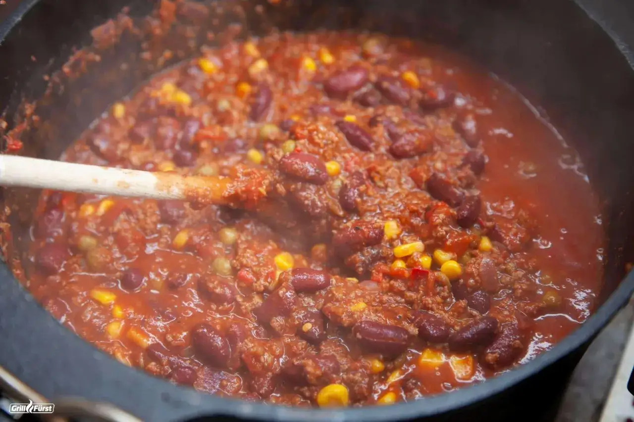Ein dampfender Topf mit Chili con Carne, voller Kidneybohnen, Hackfleisch und Mais. Ein Holzlöffel rührt in der herzhaften Mischung.