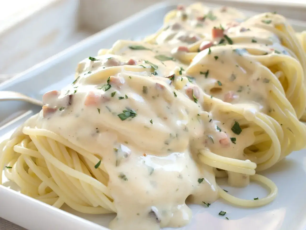 Cremige Spaghetti Carbonara mit Speckst&uuml;ckchen und Petersilie, serviert auf einem wei&szlig;en Teller.