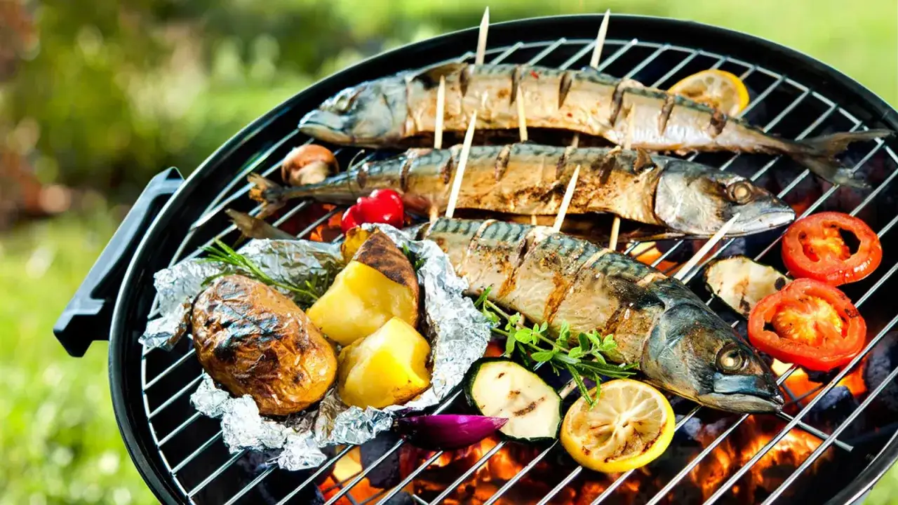Drei ganze Makrelen werden auf einem Grill zubereitet, zusammen mit Kartoffeln in Alufolie, Tomaten, Zucchini und Zitronenscheiben. Das Makrele grillen ist fast fertig.