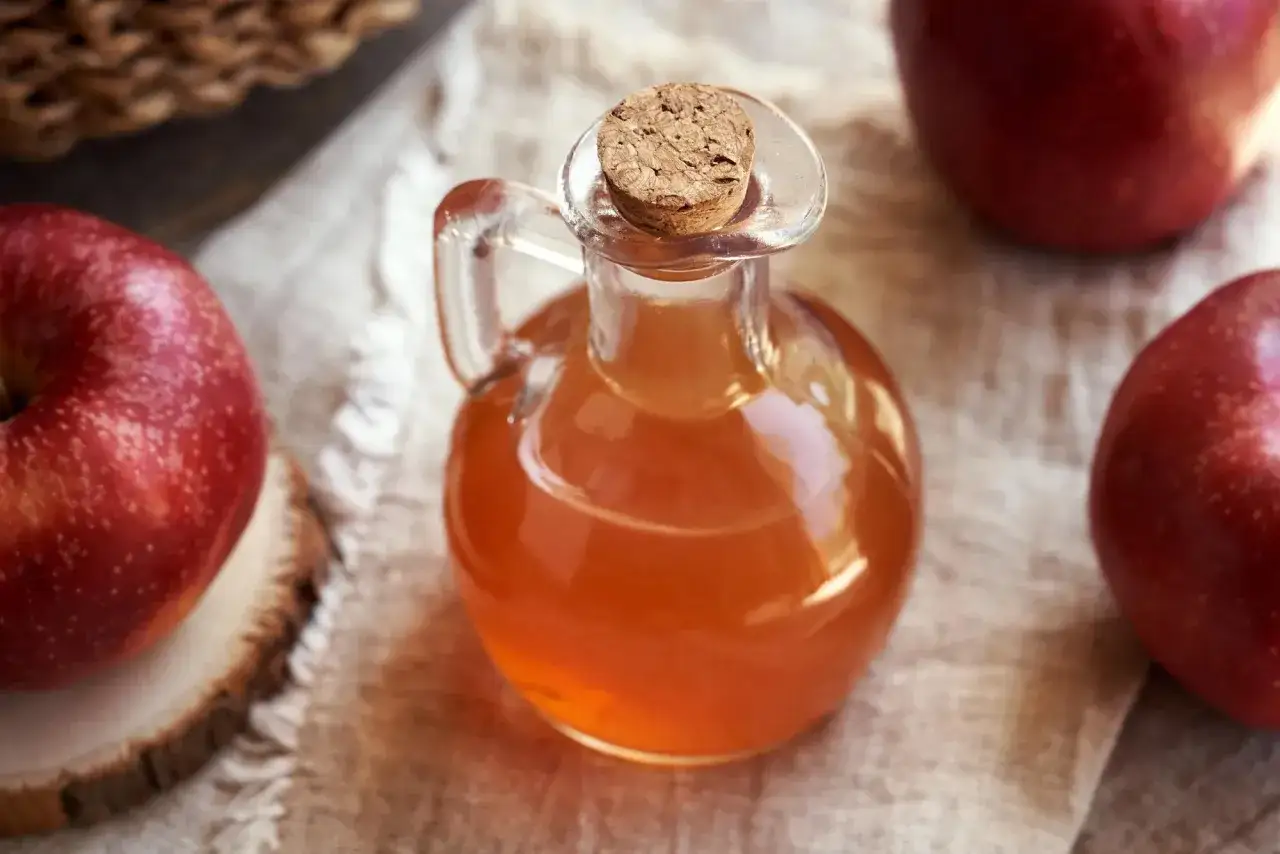 Kleine Glasflasche mit bernsteinfarbenem Apfelessig, umgeben von roten &Auml;pfeln. Ein gesunder Start in den Tag: Apfelessig trinken!