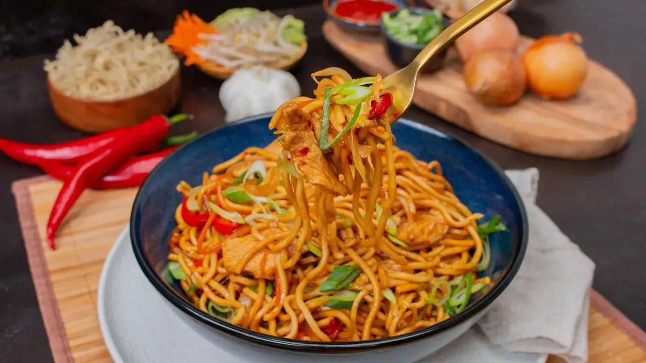 Eine Gabel hebt Nudeln mit H&uuml;hnchen und Gem&uuml;se aus einer Sch&uuml;ssel Bami Goreng. Zutaten wie Chilis und Zwiebeln sind im Hintergrund zu sehen.
