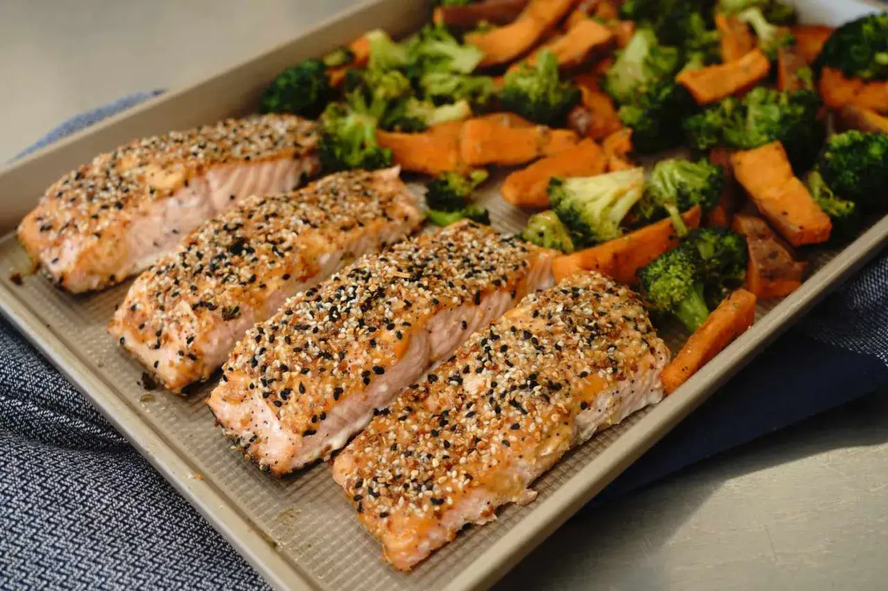 Vier Lachsfilets mit Sesamkruste auf einem Blech mit geröstetem Brokkoli und Süßkartoffeln. Perfekt für eine pescetarier-Ernährung, die Fisch erlaubt.