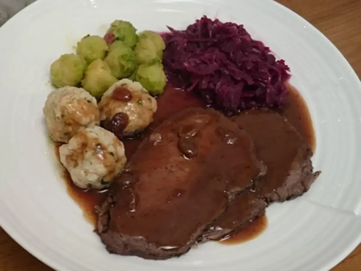 Ein Teller mit Scheiben von Sauerbraten, Klößen, Rosenkohl und Rotkohl. Ein klassisches Sauerbraten Rezept.