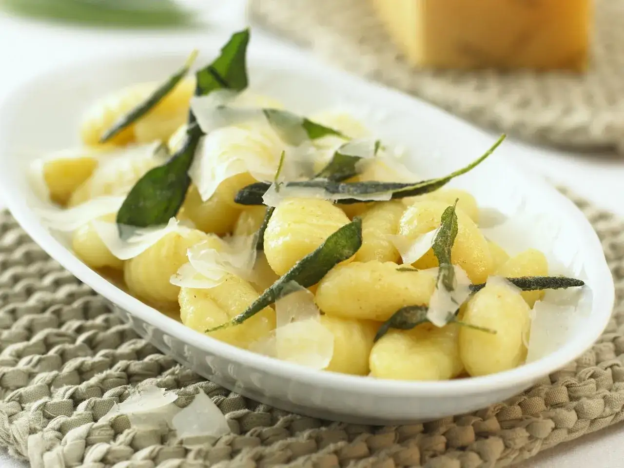 Salbei Gnocchi mit Parmesanflocken und frischen Salbeibl&auml;ttern in einer wei&szlig;en Schale auf einem geflochtenen Untersetzer.